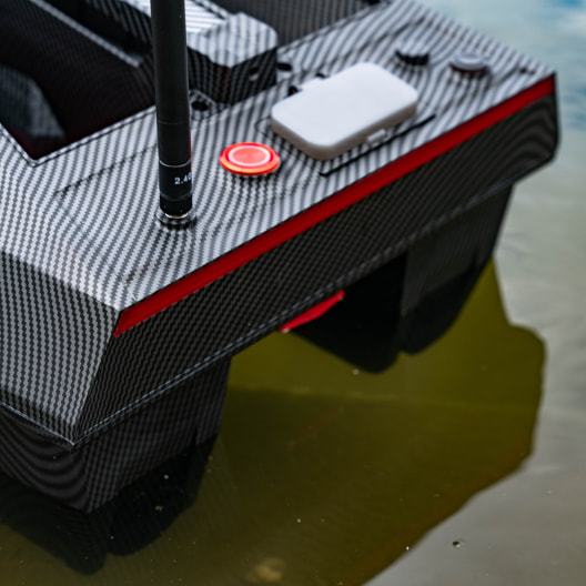 Close-up detail of a high-end carbon fiber GPS bait boat with red accent trim, showing sleek design and control interface for precision carp fishing