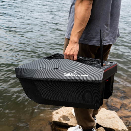 A person holding a sleek black modern fishing carbon fiber GPS bait boat with Catch Innovations branding near water's edge