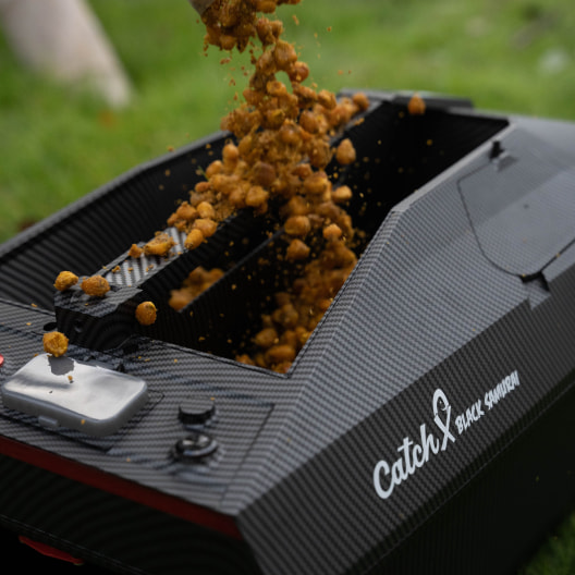 Carbon fiber CatchX bait boat hopper in action, showing bait being poured into the loading compartment in a lake background